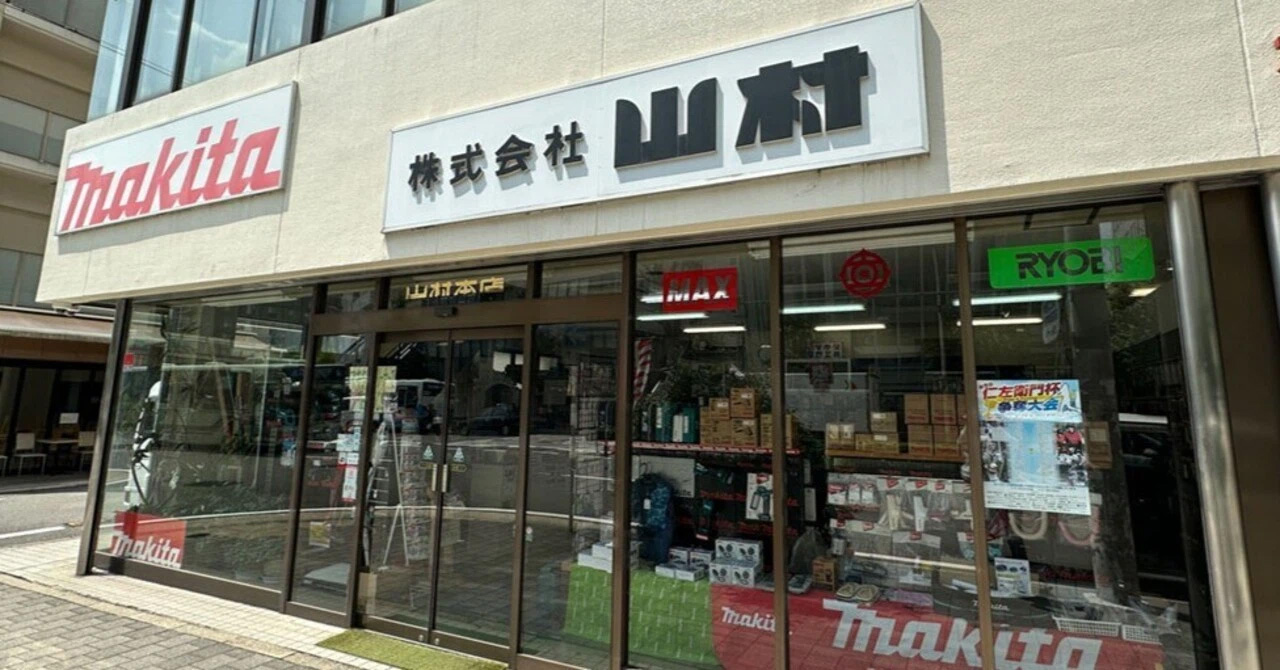 「株式会社山本」という会社の店先の写真。建物の正面には「株式会社 山本」という看板があり、その左側には「Makita」、右側には「RYOBI」のロゴも見えます。ガラス張りの店内には、工具らしき商品が陳列されているのがわかります。